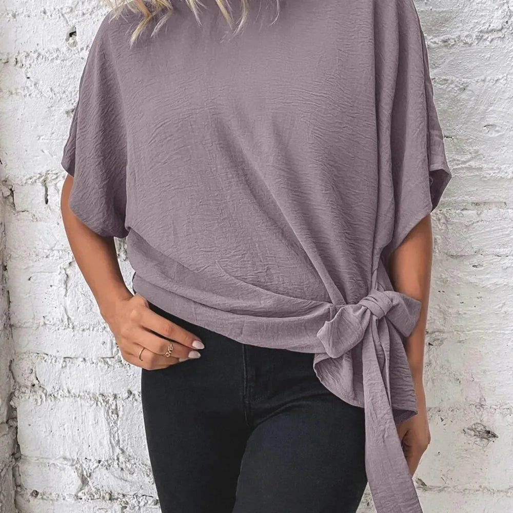 Knotted Round Neck Half Sleeve Blouse in mauve color featuring a tie detail, paired with black pants against a brick wall.