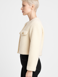 Side view of a woman wearing a Faux Shearling Button-Front Cropped Jacket in cream color, showcasing its chic design.