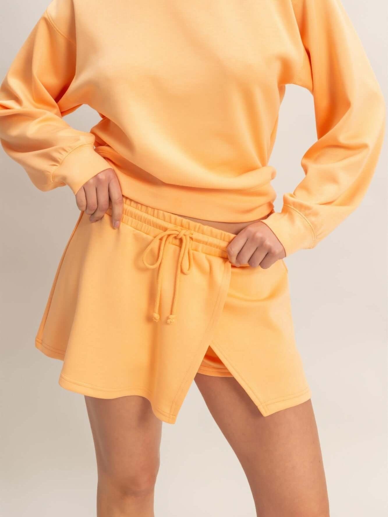Model wearing a Wrap Front Elastic Waist Skort in soft orange fabric with a matching oversized sweatshirt.