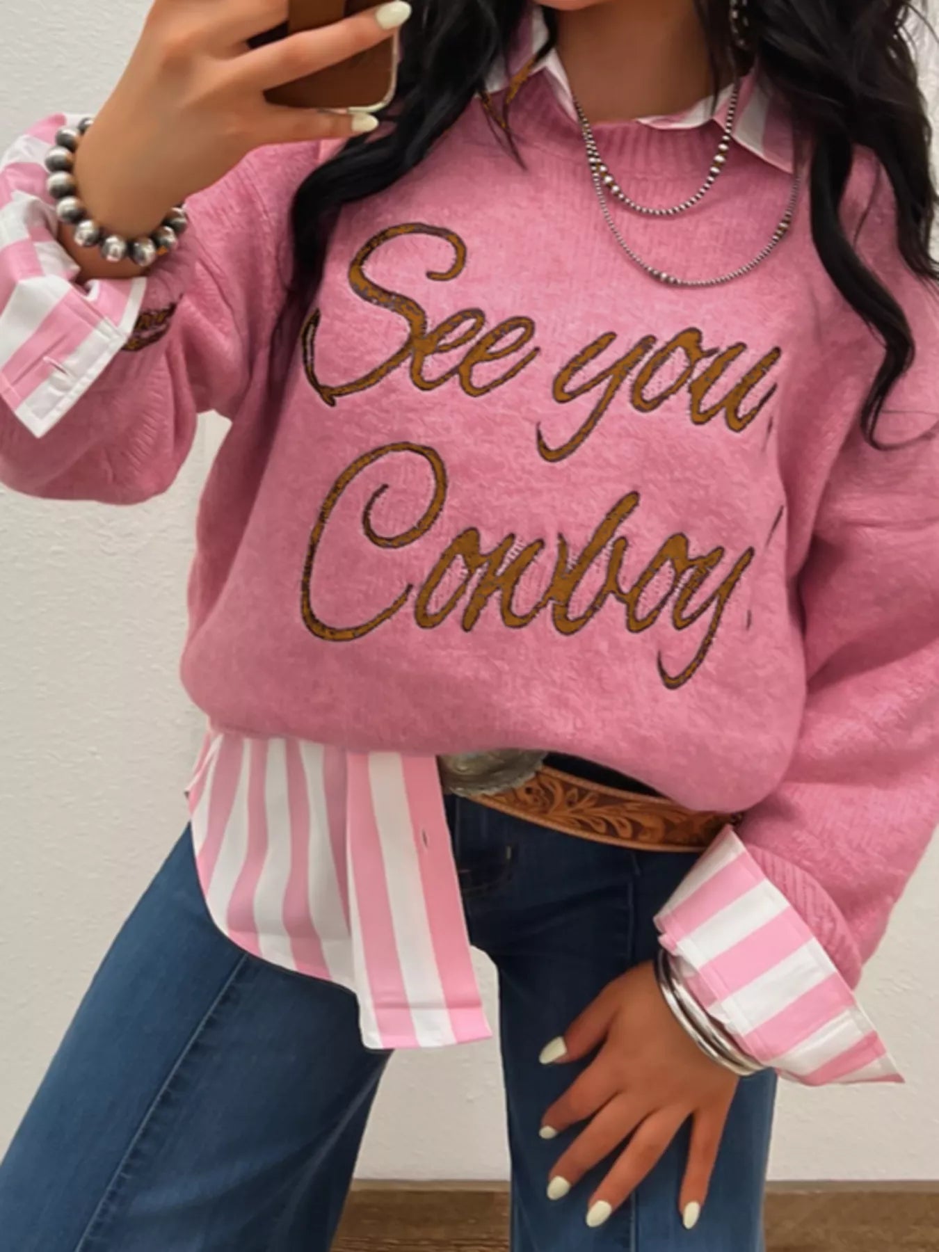Woman wearing a pink See You Cowboy Round Neck Sweater styled with a striped shirt and denim jeans.