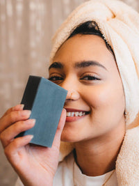 Smiling woman with towel holding Keika Charcoal Black Soap Bar, promoting skincare for oily skin and eczema.