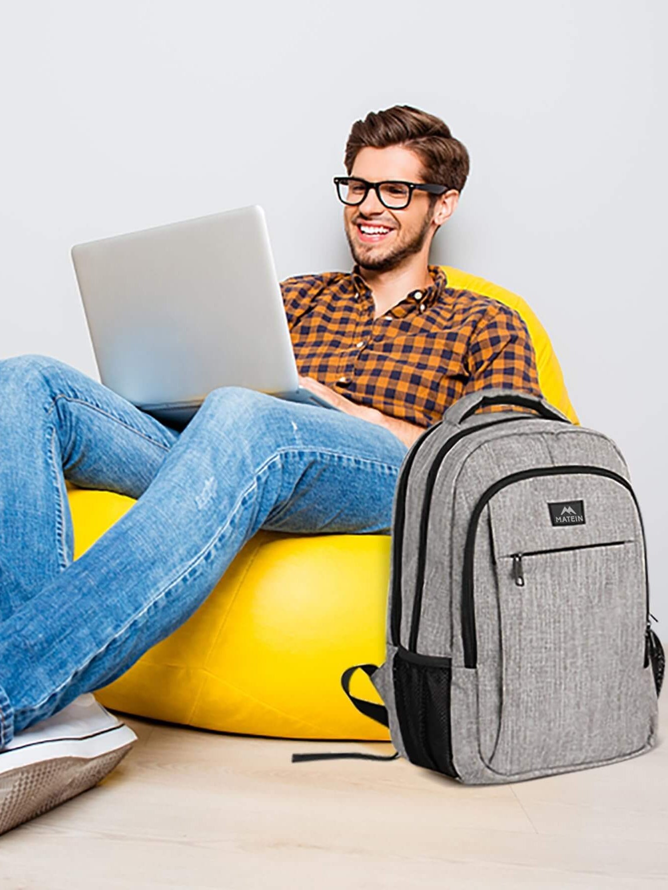 Man sitting on a yellow bean bag using a laptop with a MATEIN travel laptop backpack nearby.