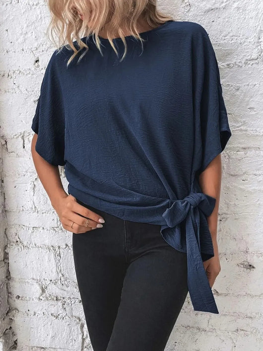 Knotted Round Neck Half Sleeve Blouse in navy blue with tied detail, styled with black pants against a textured wall.