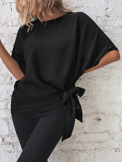 Knotted Round Neck Half Sleeve Blouse in black showcased against a textured wall, featuring a stylish tied detail.