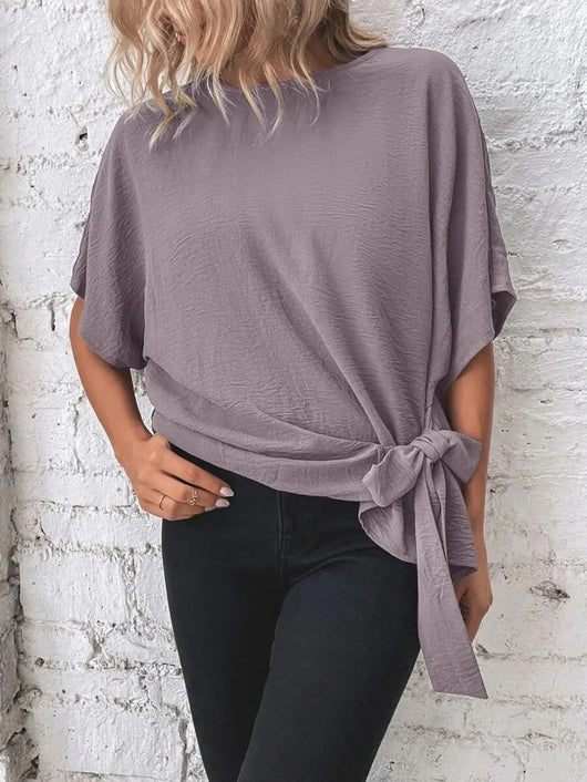 Knotted Round Neck Half Sleeve Blouse in mauve color featuring a tie detail, paired with black pants against a brick wall.
