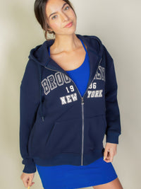 Model wearing a navy Graphic Drop Shoulder Zipper Up Hoodie with Brooklyn logo over a blue dress. Stylish and comfortable casual wear.