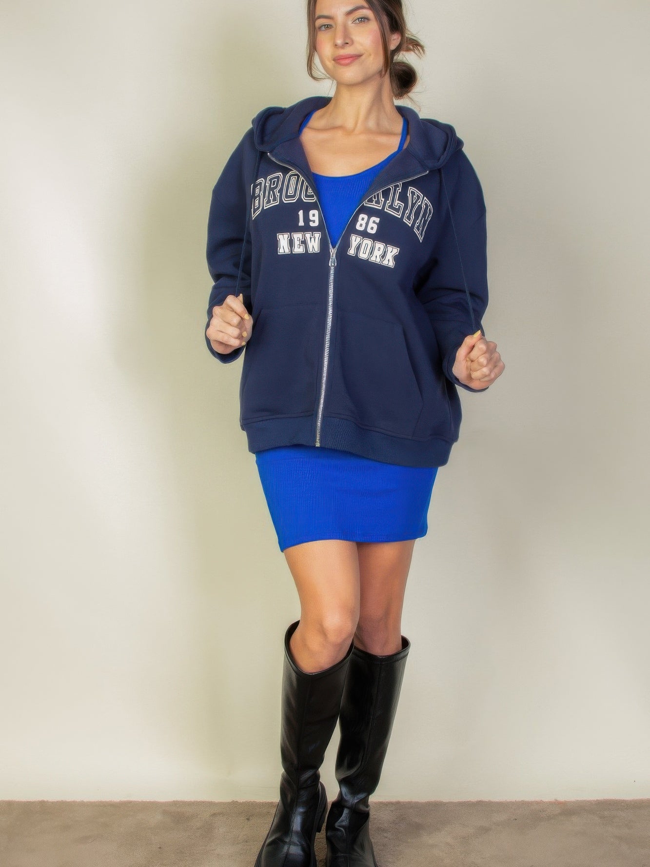 Model wearing a Graphic Drop Shoulder Zipper Up Hoodie with Brooklyn logo, styled with a blue dress and knee-high boots.