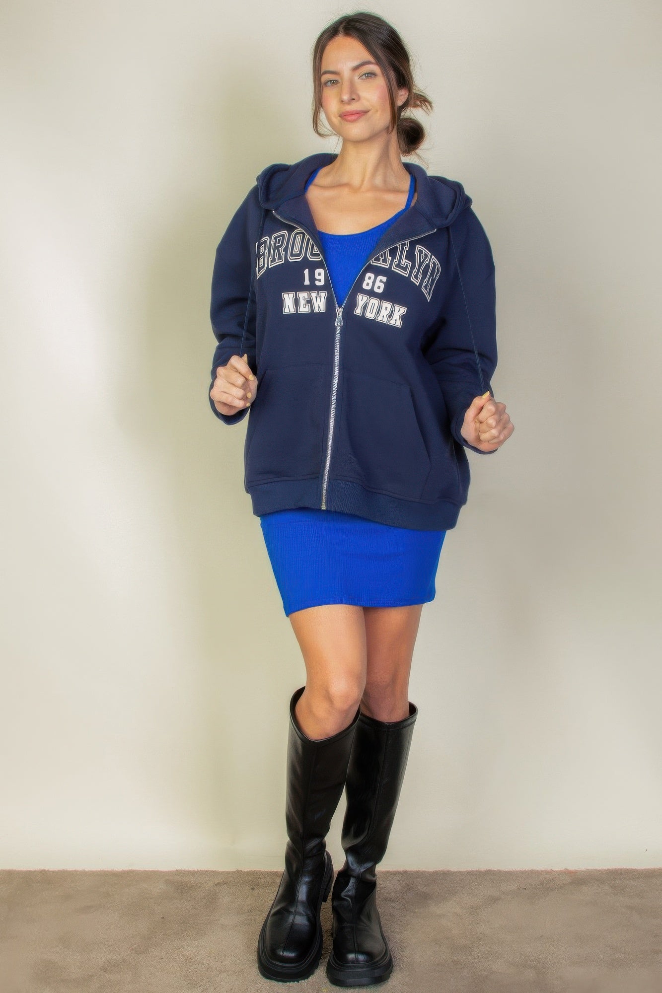 Model wearing a Graphic Drop Shoulder Zipper Up Hoodie with Brooklyn logo, styled with a blue dress and knee-high boots.