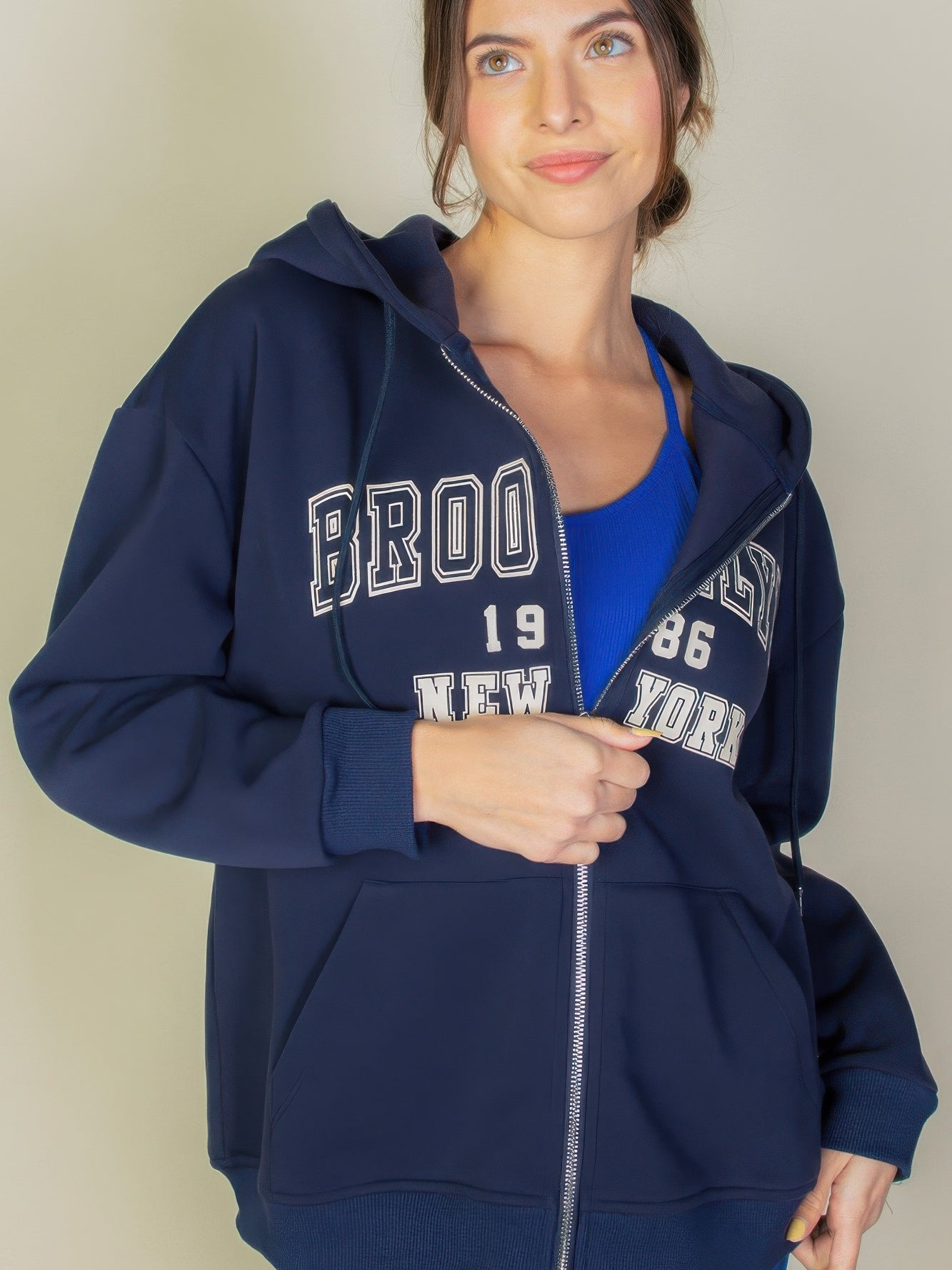 Woman wearing a navy Graphic Drop Shoulder Zipper Up Hoodie with Brooklyn logo and front pockets.