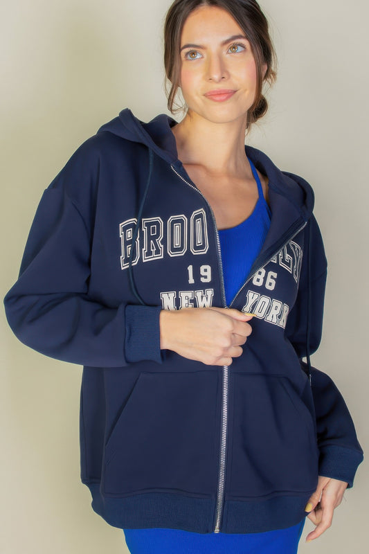 Woman wearing a navy Graphic Drop Shoulder Zipper Up Hoodie with Brooklyn logo and front pockets.