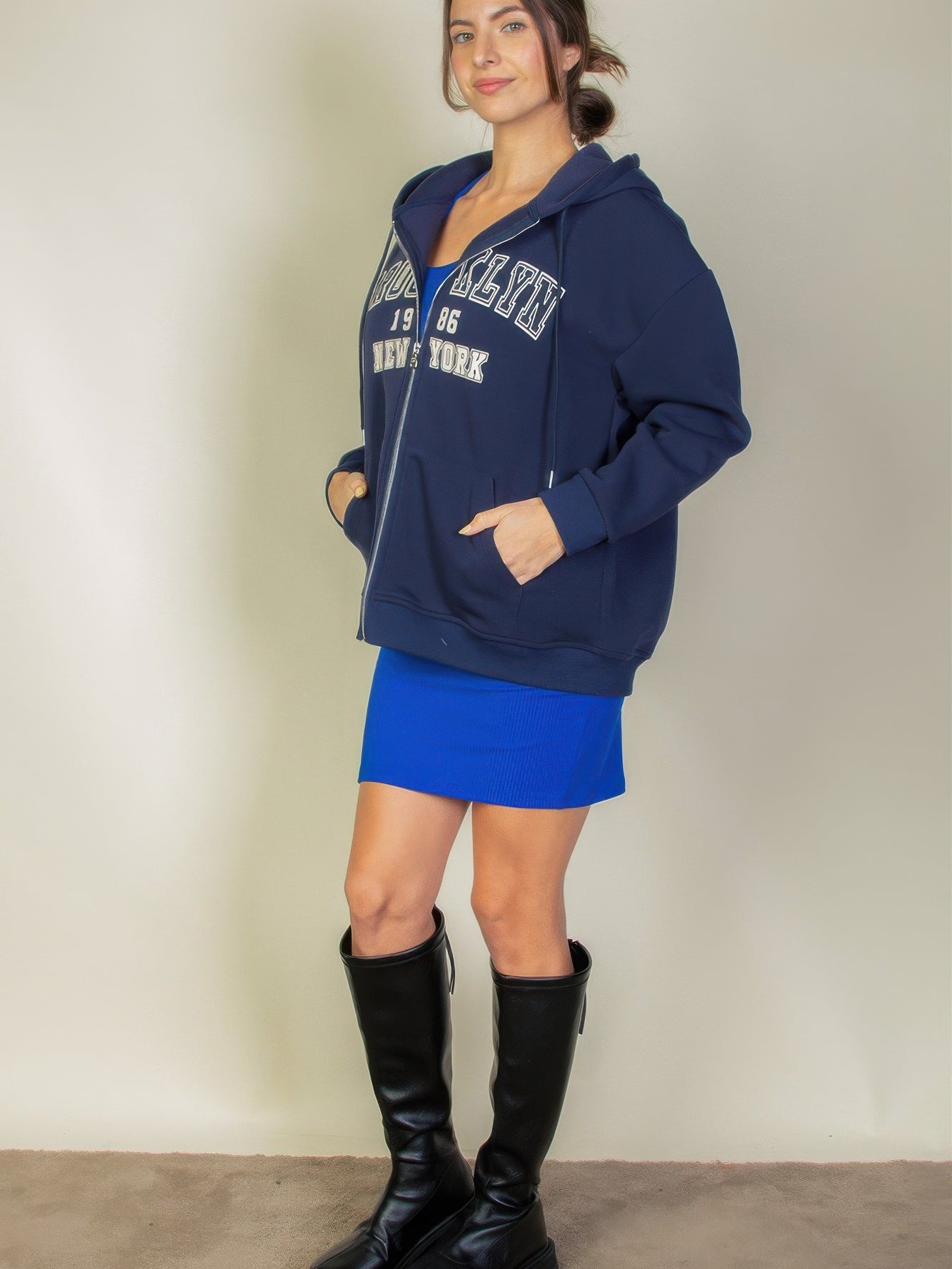 Woman modeling a navy graphic drop shoulder zipper up hoodie with Brooklyn logo, paired with a blue skirt and black knee-high boots.