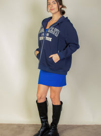 Woman modeling a navy graphic drop shoulder zipper up hoodie with Brooklyn logo, paired with a blue skirt and black knee-high boots.