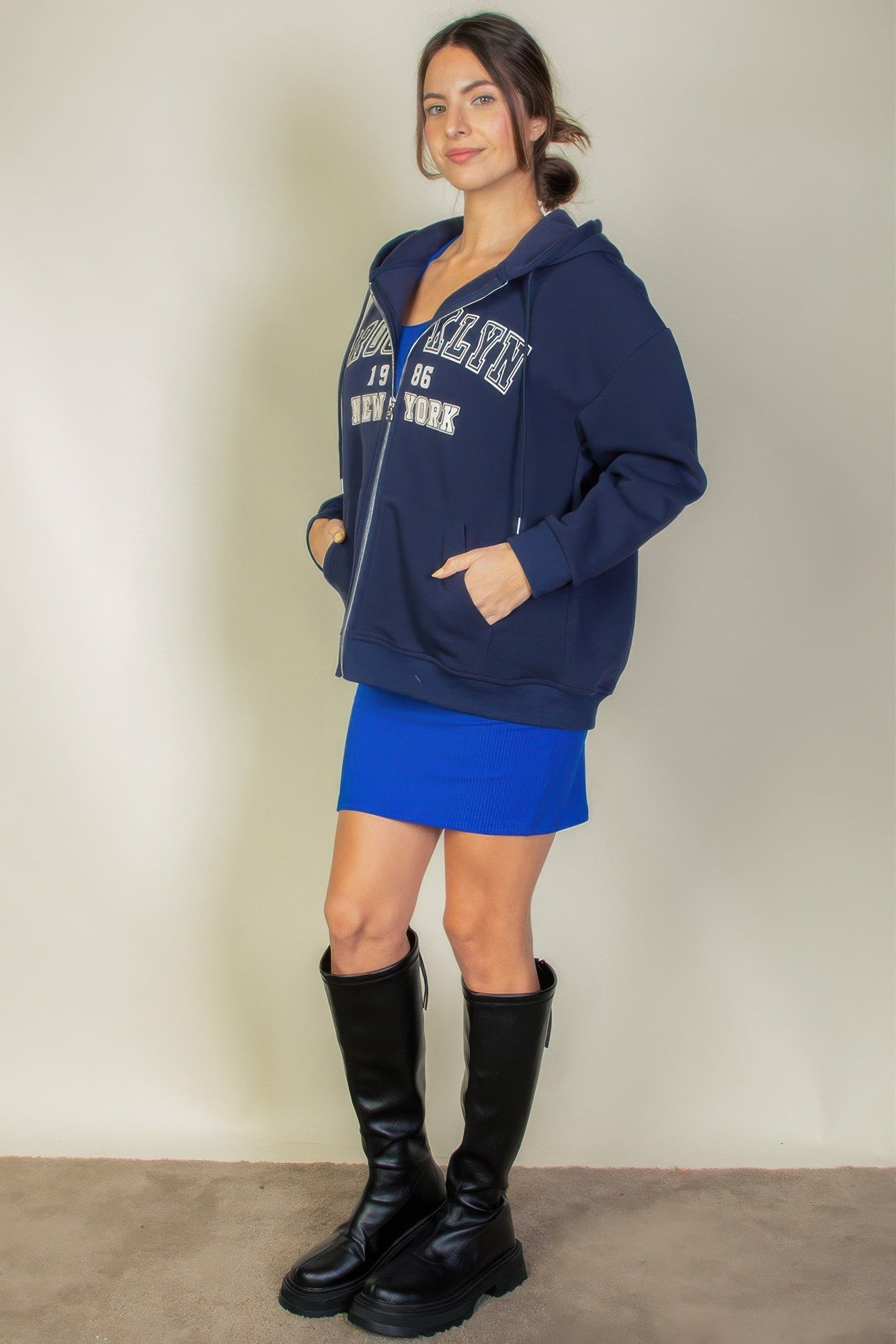 Woman modeling a navy graphic drop shoulder zipper up hoodie with Brooklyn logo, paired with a blue skirt and black knee-high boots.