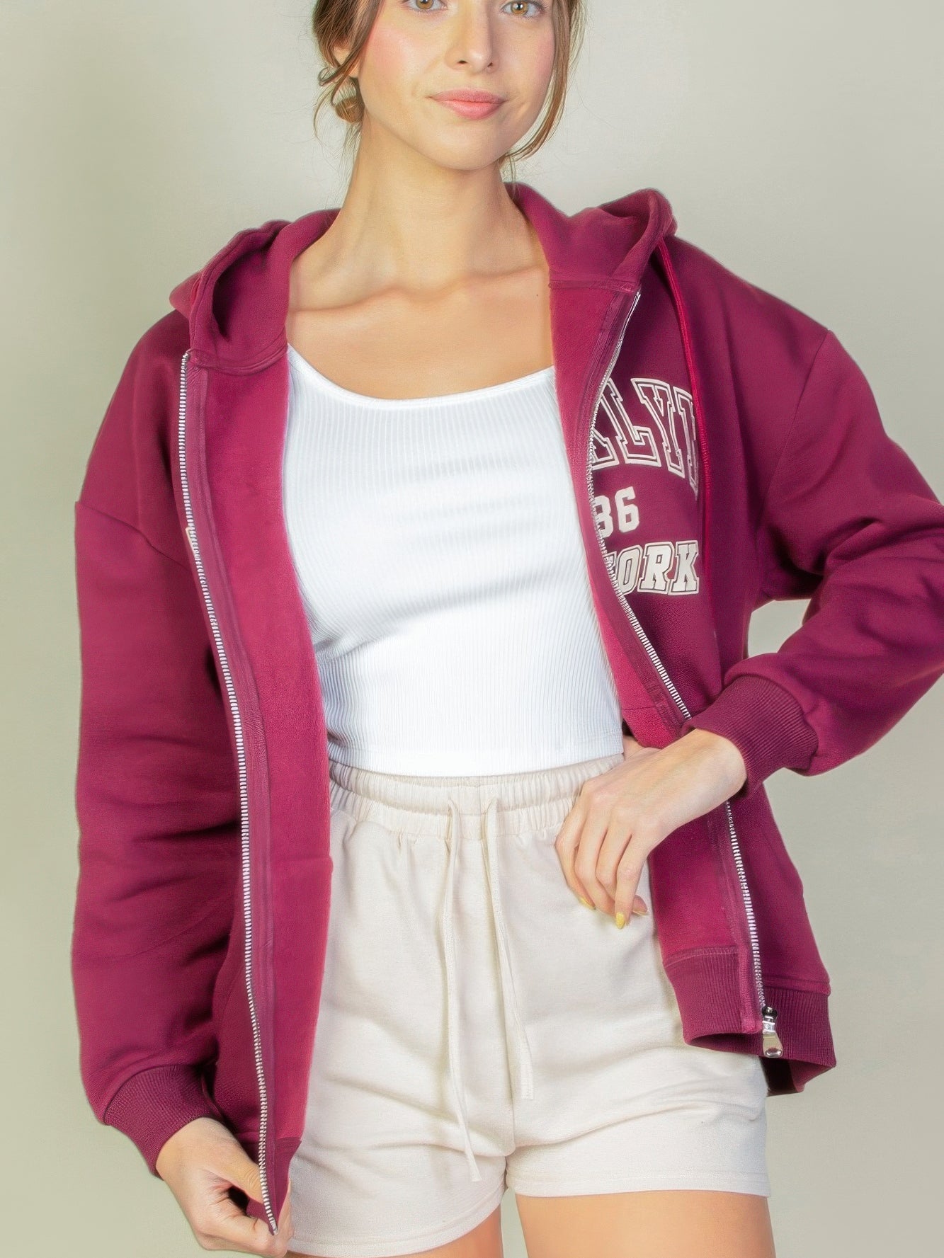 Model wearing a maroon Graphic Drop Shoulder Zipper Up Hoodie over a white tank top and beige shorts.