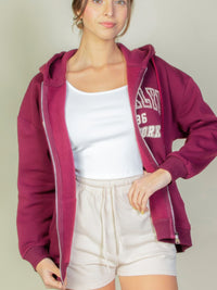 Model wearing a maroon Graphic Drop Shoulder Zipper Up Hoodie over a white tank top and beige shorts.