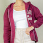 Model wearing a maroon Graphic Drop Shoulder Zipper Up Hoodie over a white tank top and beige shorts.