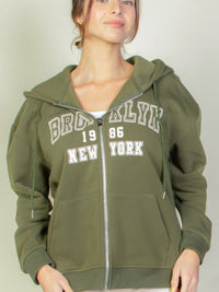 Model wearing a graphic drop shoulder zipper up hoodie in olive green with Brooklyn logo, perfect for casual style.