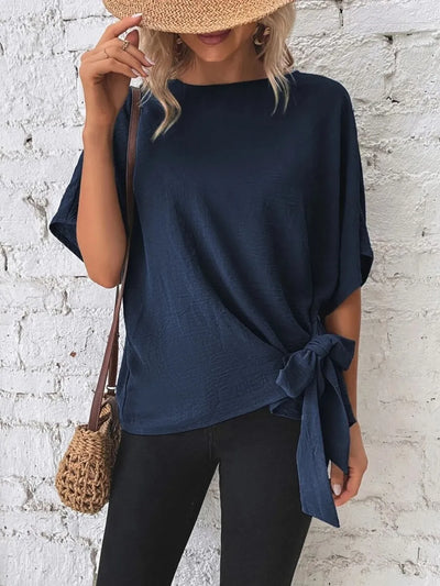 Knotted Round Neck Half Sleeve Blouse in navy worn with a straw hat and stylish bag against a white brick wall.