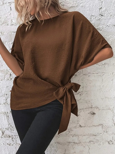 Knotted Round Neck Half Sleeve Blouse styled with a side tie, showcasing a cozy brown color against a brick wall.